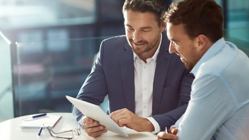 Two businessmen in a bright office review documents at a round table, with glasses and notebooks nearby.