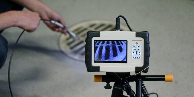 Portable inspection camera on a tripod displaying a metal drain grate on its screen, viewed up close