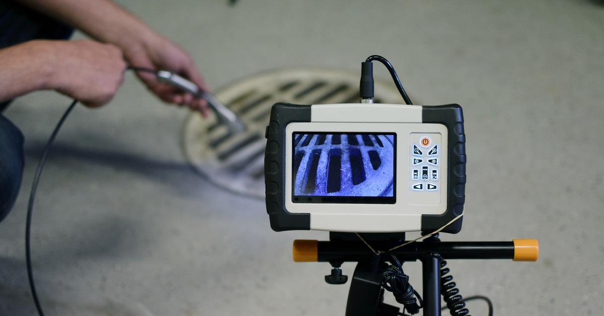 Portable inspection camera on a tripod displaying a metal drain grate on its screen, viewed up close